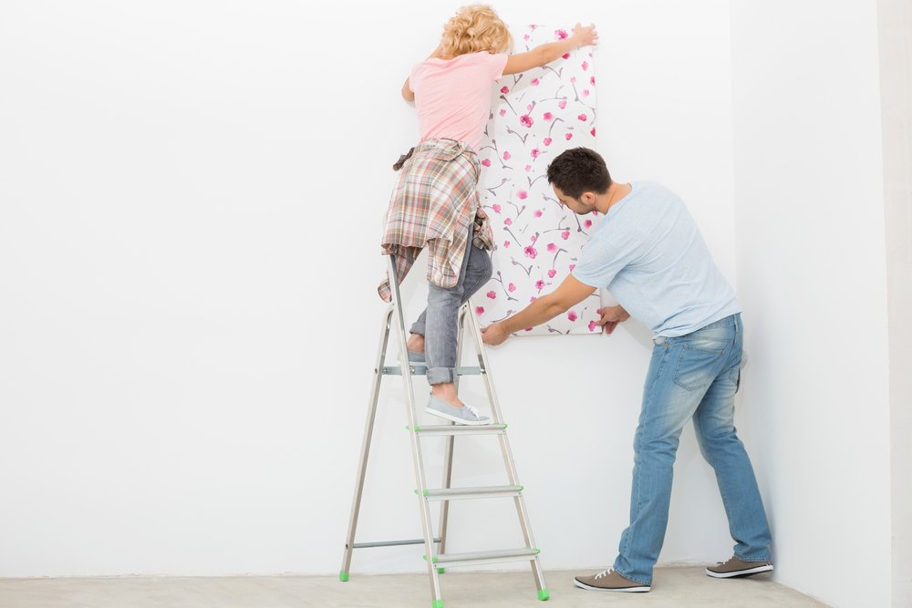 Een persoon brengt renovlies van 150 gram aan op een muur met een roller, terwijl een andere persoon het gladstrijkt met een behangspatel.