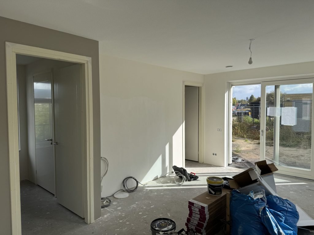 Doorkijk van een nieuwbouwwoonkamer naar de hal, met gesausde wanden en materiaal op de vloer