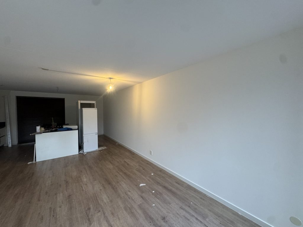 Lange witte woonkamerwand waar het licht schuin langs valt, met een houten vloer en een keukenblok achterin
