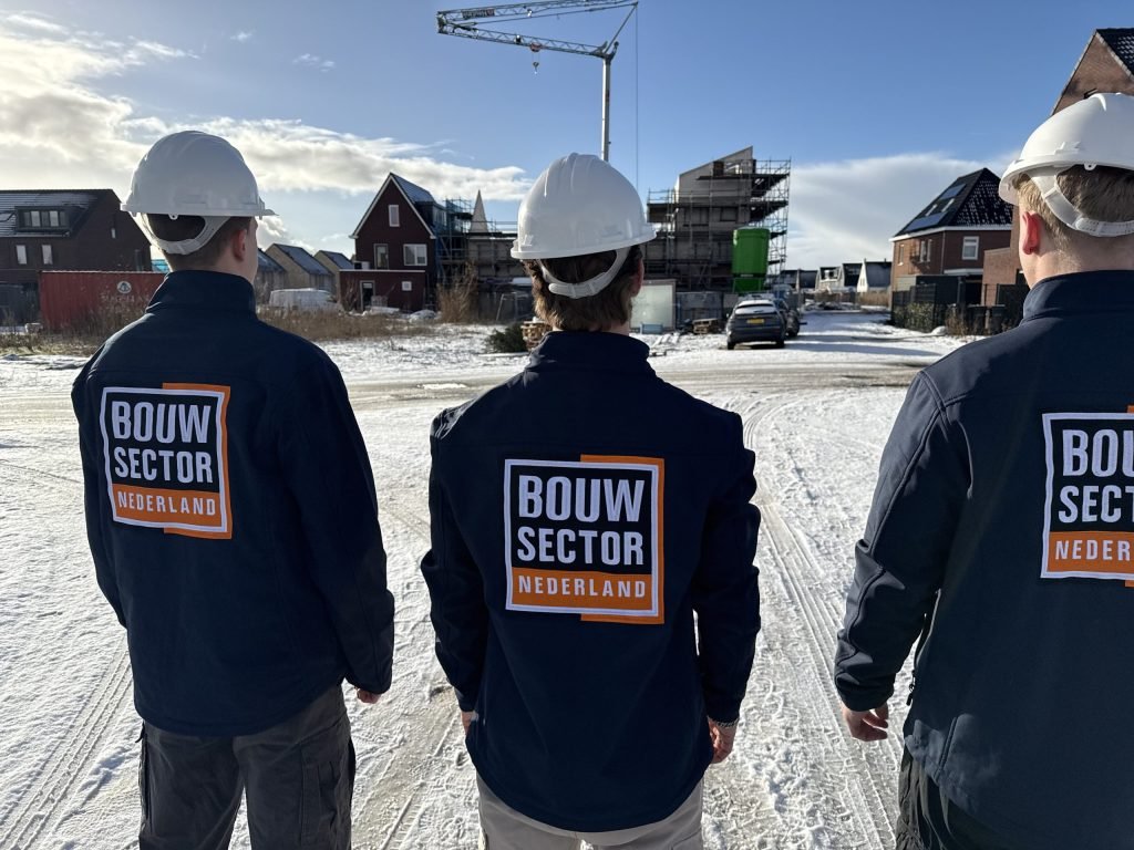 Drie vakmensen van Bouwsector Nederland met witte helm en logo op de rug in een besneeuwde nieuwbouwwijk